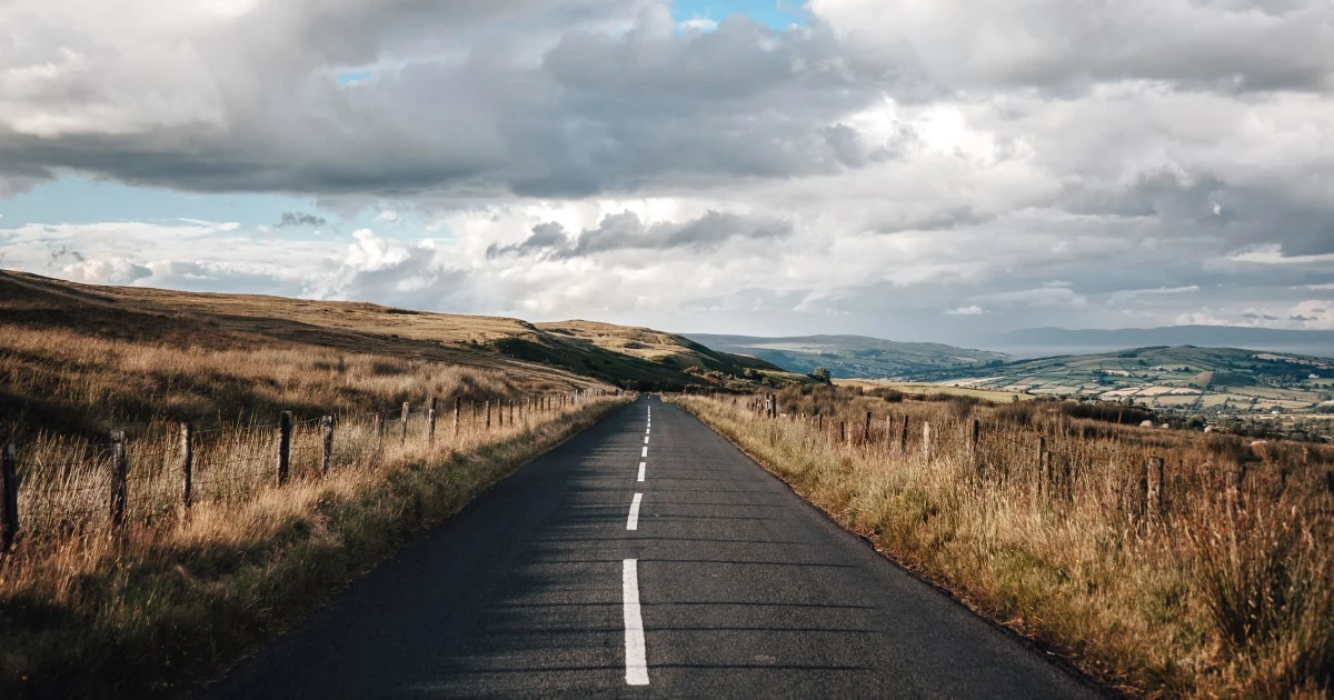 Empty two-lane road winding through golden grassland hills under a cloudy sky.