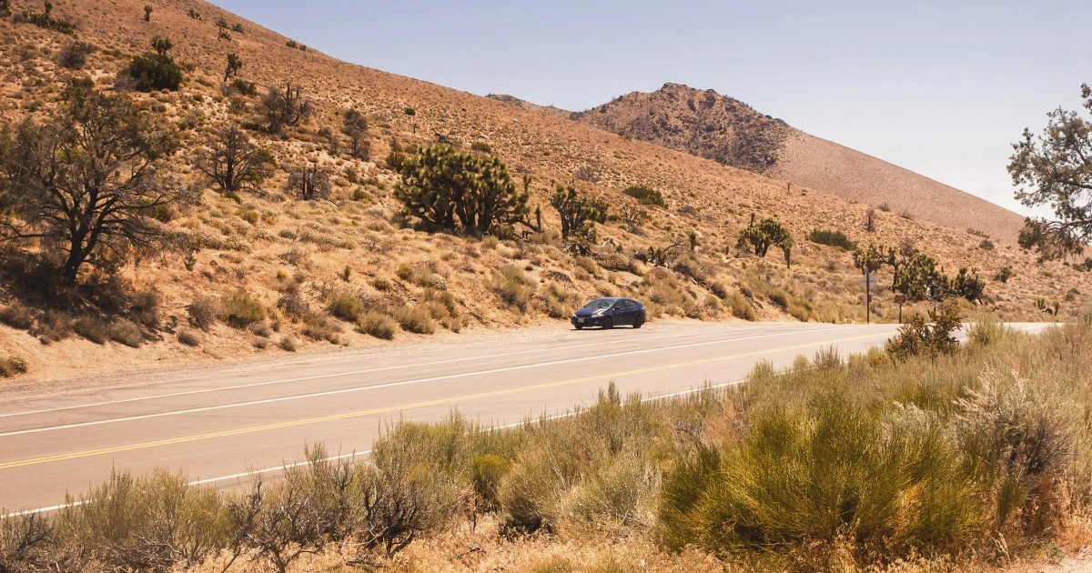 Dark car driving along a road through dry, sun-baked desert hills.
