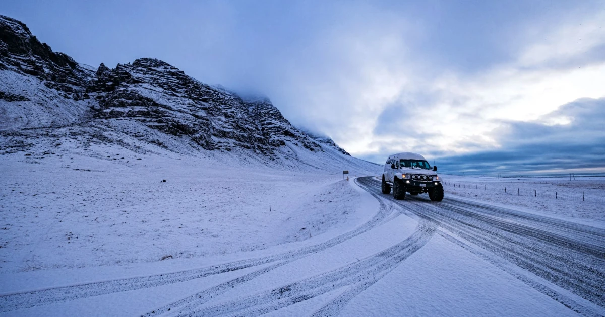 SUV driving on a snow-covered road toward snowy mountains under a blue sky.