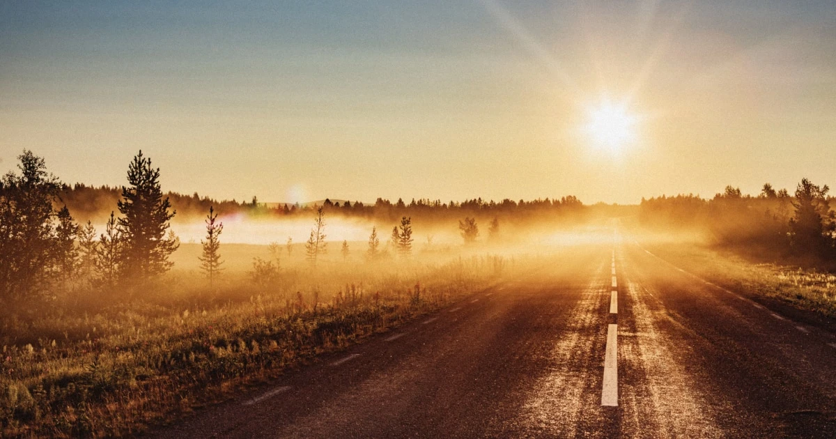 Straight road at sunrise, with the sun low over misty fields and warm golden light.