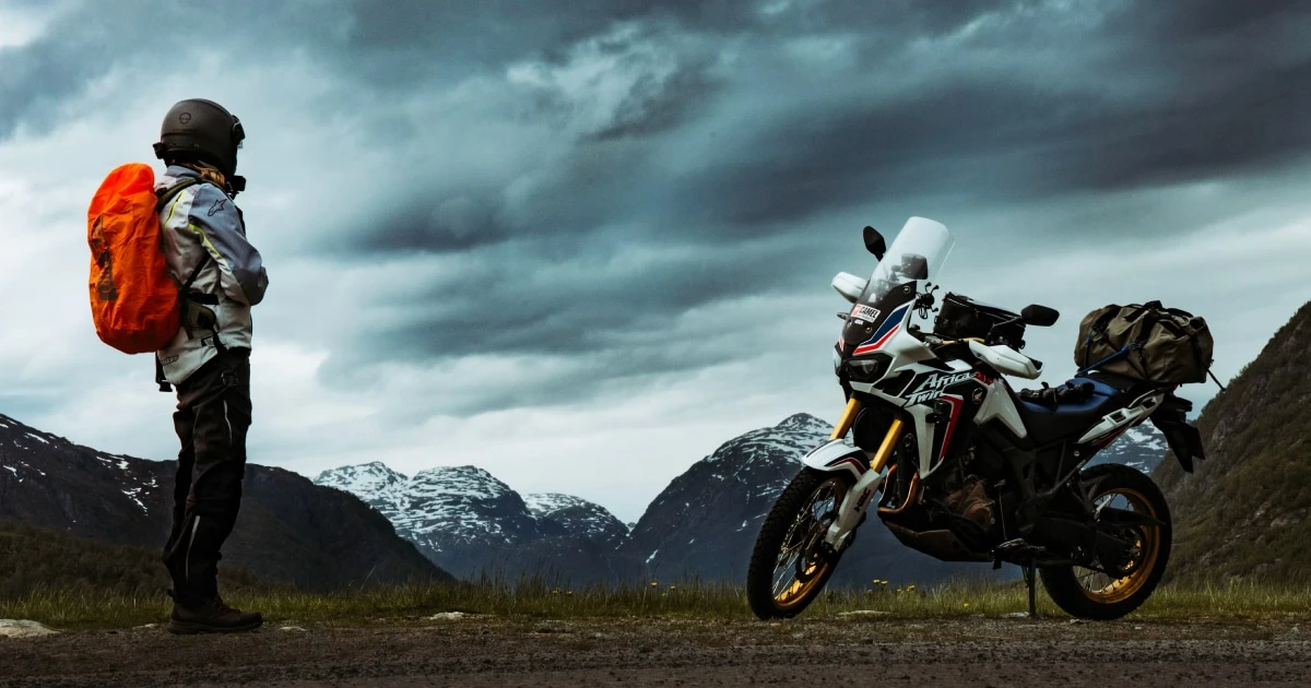 Rider with an orange backpack standing beside an adventure motorcycle in the mountains under a stormy sky.