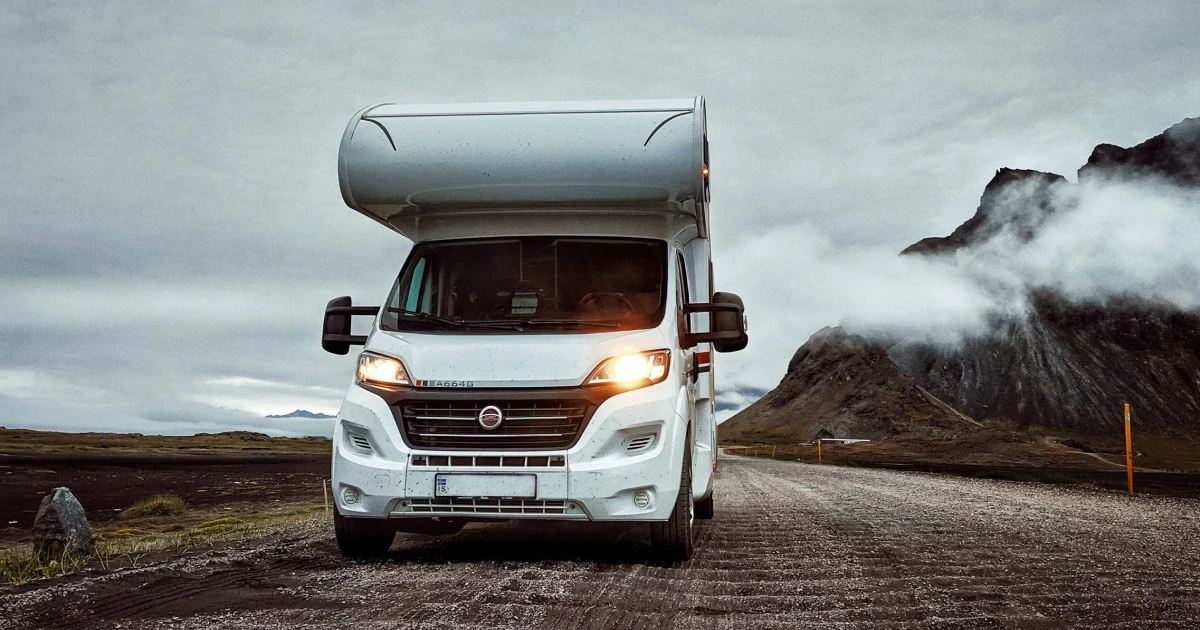 White motorhome with its headlights on, parked on a mountain road with clouds around the peaks.