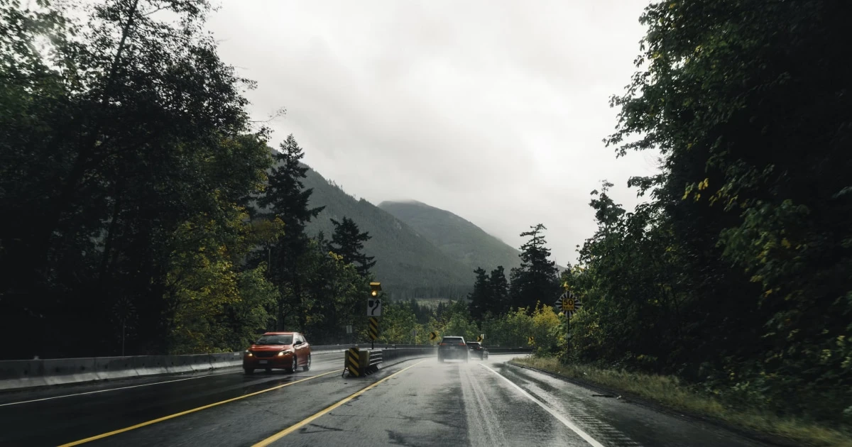 Cars driving on a wet road through forested mountains under an overcast sky.