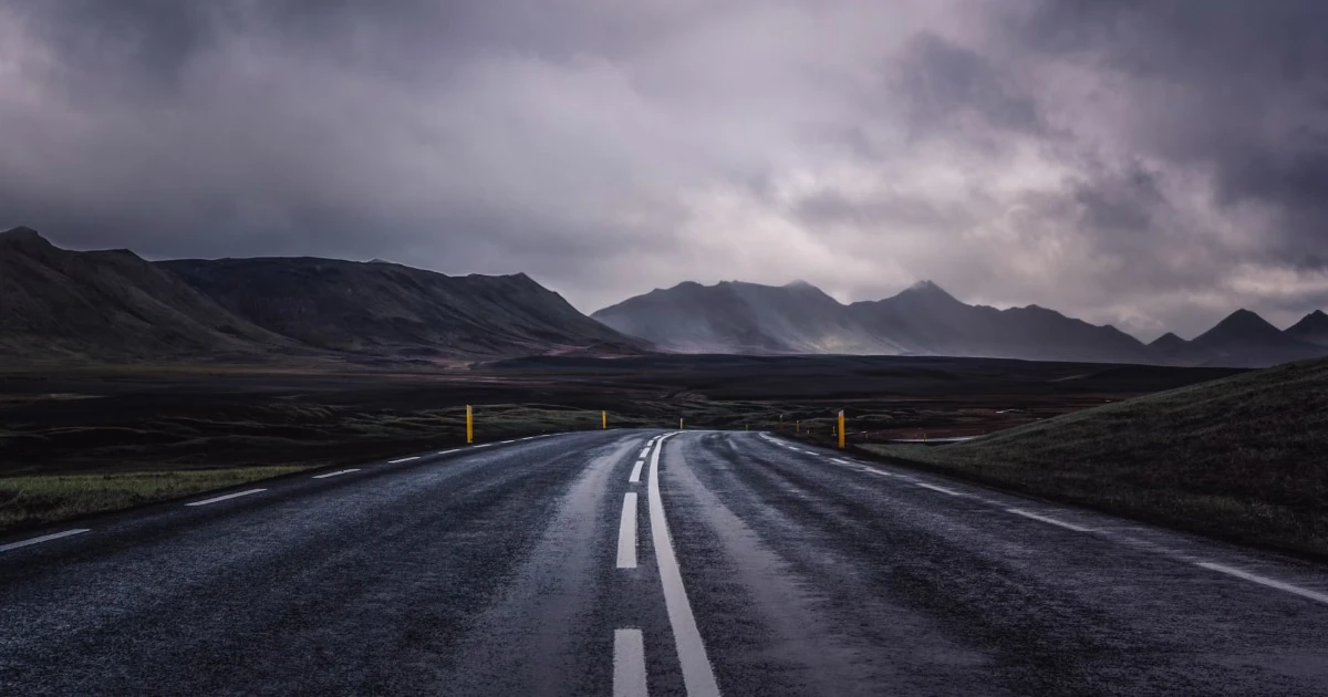 Wet road reflecting the sky, heading toward mountains under dark rain clouds.