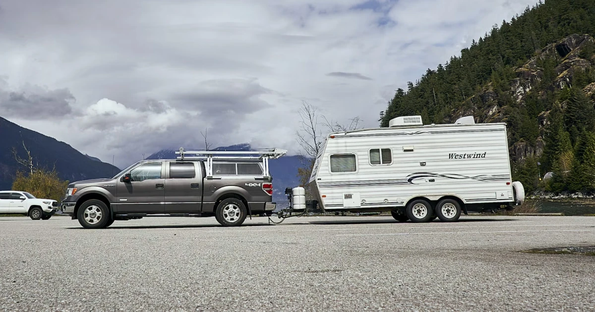 Pickup truck parked next to a travel trailer below a forested mountain on a cloudy day.