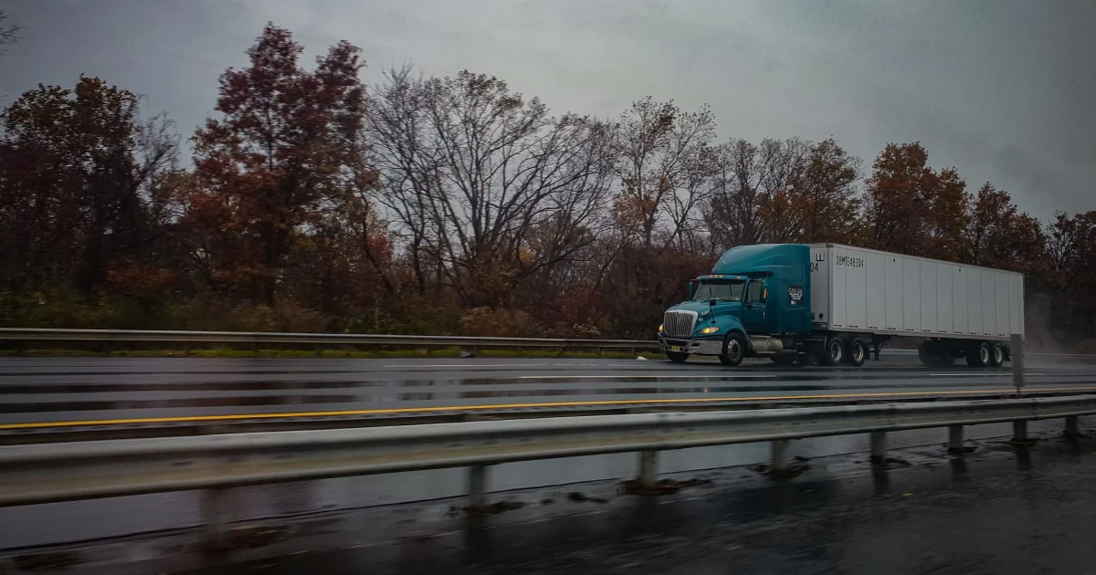Semi-truck with a teal cab driving on a wet highway beside a guardrail under a gray autumn sky.