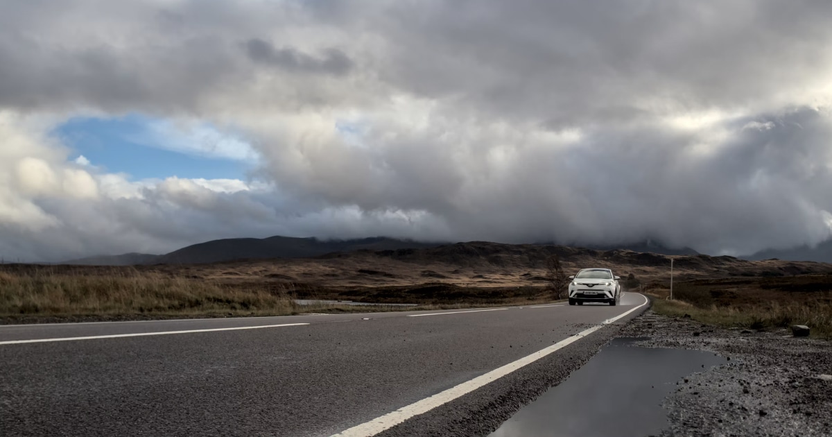 Lone car on a curving road heading toward dark storm clouds, with a patch of blue sky above.