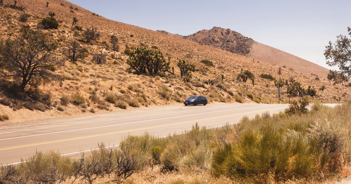 Dark car driving along a road through dry, sun-baked desert hills.