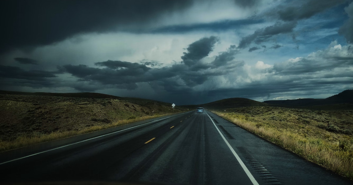 Empty road stretching beneath dark, heavy storm clouds over green hills.