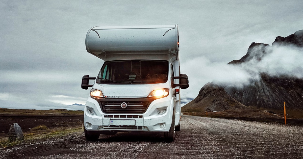 White motorhome with its headlights on, parked on a mountain road with clouds around the peaks.