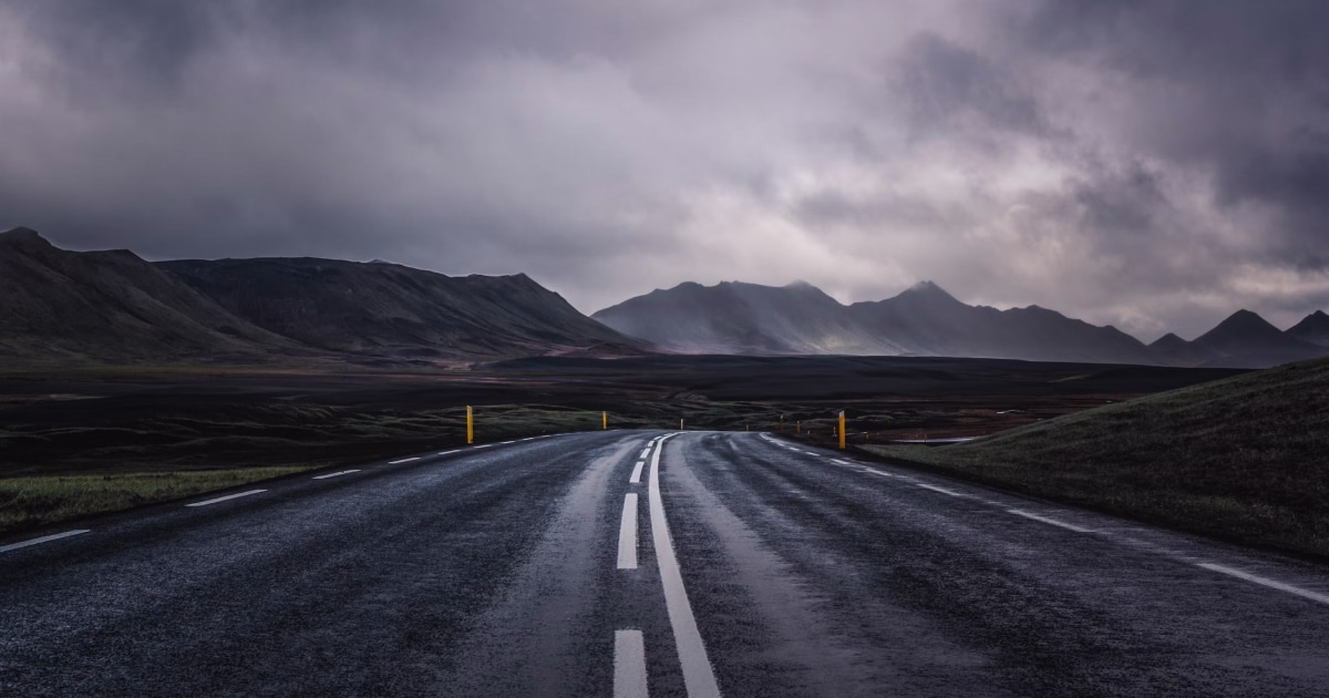 Wet road reflecting the sky, heading toward mountains under dark rain clouds.