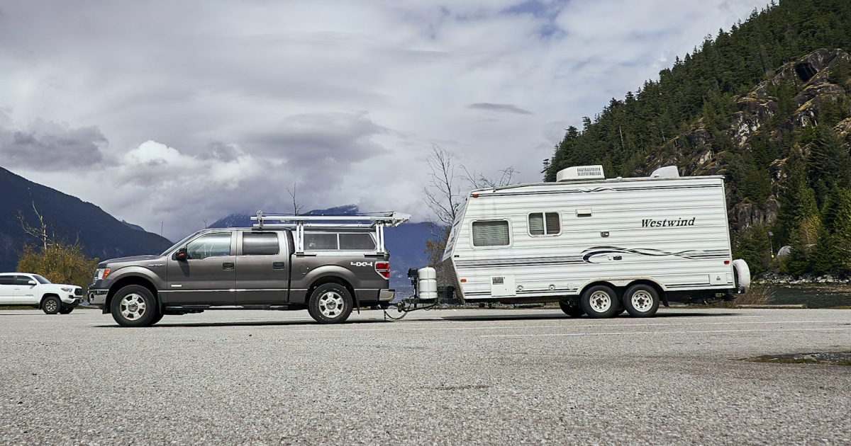 Pickup truck parked next to a travel trailer below a forested mountain on a cloudy day.