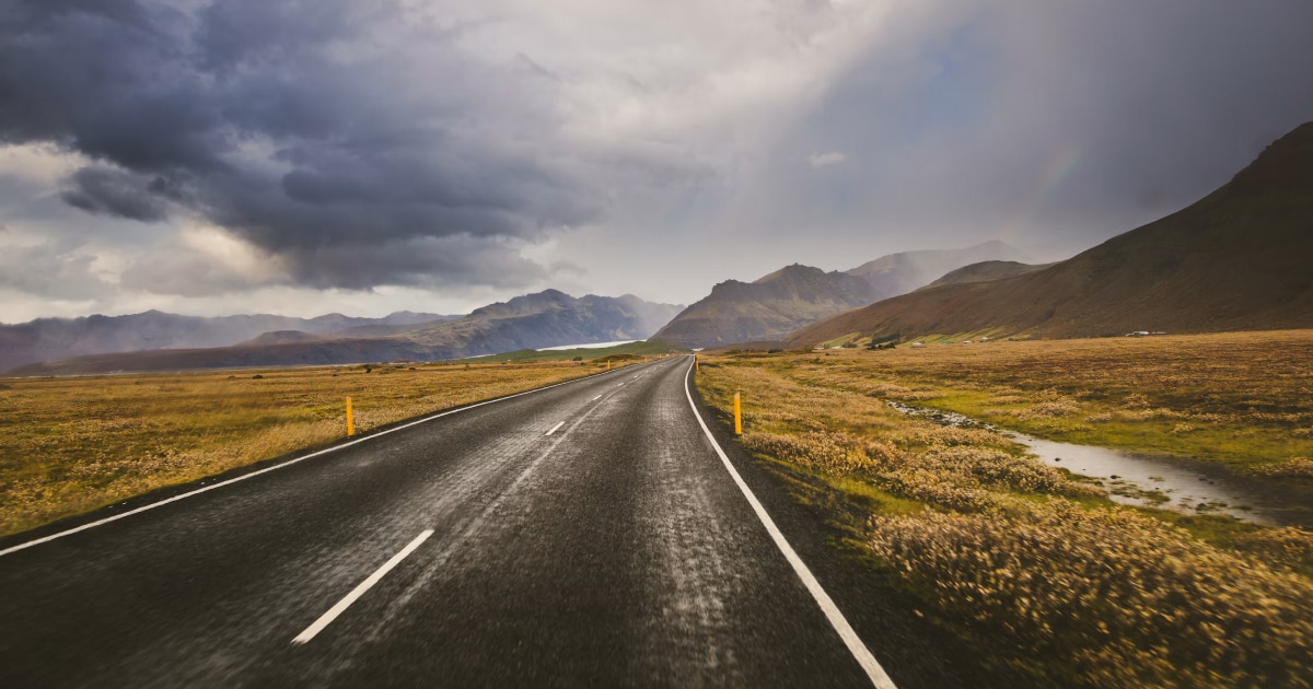 Wet road curving through golden grassland toward mountains under a moody, changing sky.